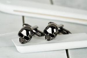 An image of two skull gun metal cufflinks facing the left side. The pair of cufflinks displays a DM, Brooklyn etched hexagonal logo.