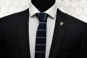 A Dear Martian, navy striped tie modeled on a mannequin wearing a suit. The tie is paired with our Carroll gold fleur de lis lapel pin.