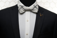 An image of a mannequin wearing both a white checked linen bow tie and gold stag lapel pin.