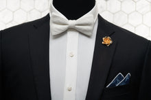 The crepe linen white bow tie is displayed on a mannequin paired with a denim pocket square and rose lapel pin.