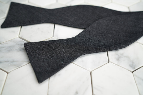 An image of Dear Martian's denim black bow tie untied, lying against a hex tiled background.