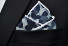A blue camo denim pocket square by Dear Martian, Brooklyn is shown folded on a black suit pocket.