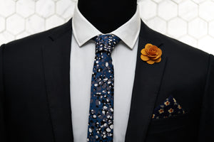 A men's suit jacket displays Dear Martian acessories, which include a floral patterned pocket square, orange flower lapel pin, and the Crooke blue steel spotted cotton tie.