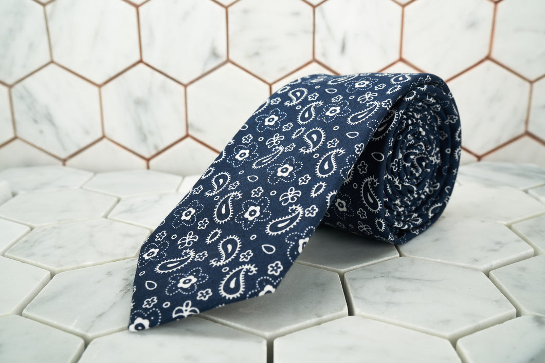 A paisley navy patterned necktie by Dear Martian, is named the Cowboy and rolled up against a white background. 