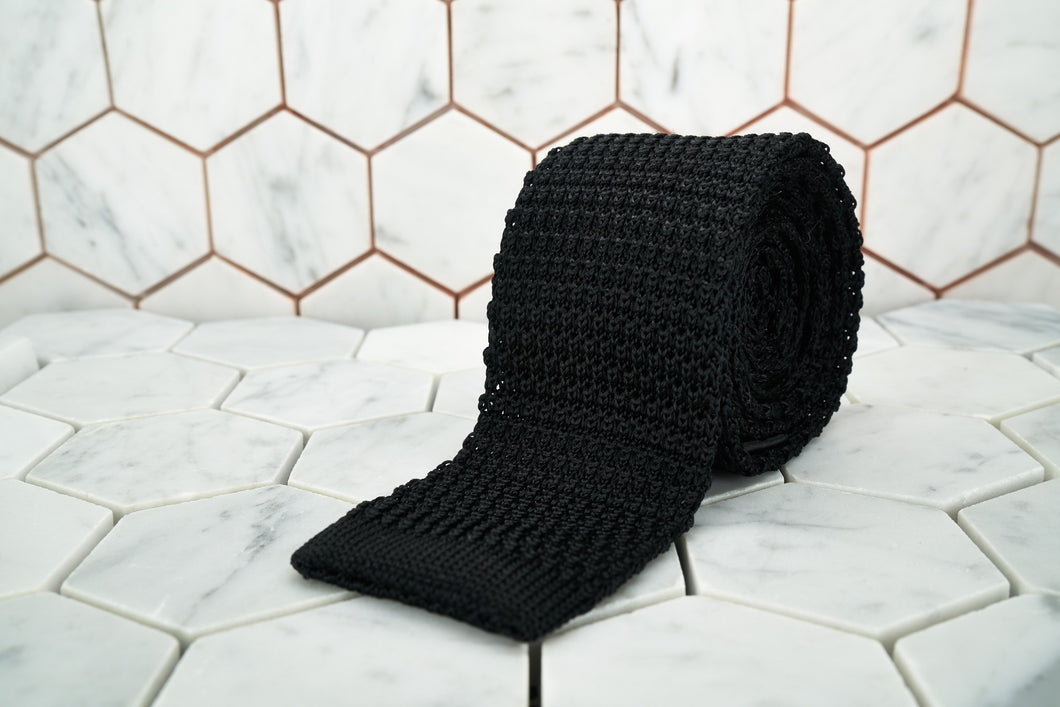 This is the Dear Martian sleek jet black, silk knitted neck tie rolled and displayed against a hexagon background.