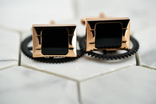 A front view image of the Carre rose gold cufflinks for men, which lay against two black cogs.