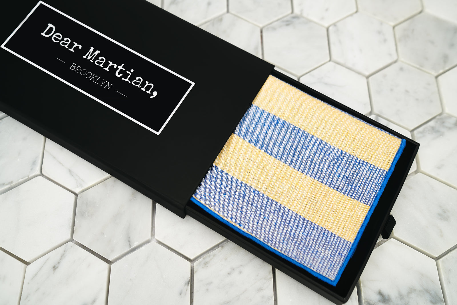 An image showing the Bookie yellow and blue striped pocketsquare. The matte black box features a pull out style with our Dear Martian, Brooklyn logo
