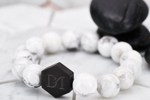 A side view image of the Jon Snow white bead bracelet by Dear Martian Brooklyn. In the background lies two black rocks for contrast of the white howlite stone.