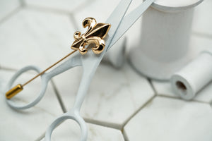 A product image of the gold fleur de lis lapel pin by Dear Martian Brooklyn, which lays upon wooden spools.