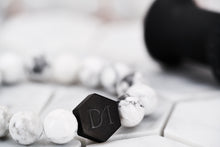 A close up image of the Dear Martian signature DM hexagon black logo bead. The bead is strung with chakra white howlite stone beads.