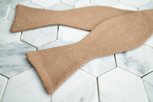 An image of a crepe-linen latte bow tie lying flat and untied against a white hexagonal background.