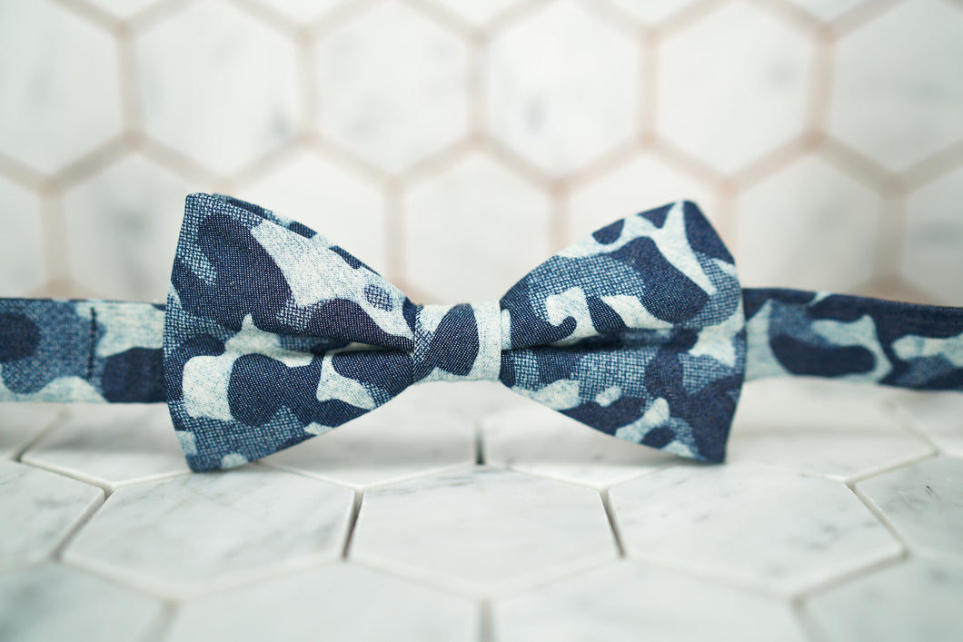 Laying against a white hexagon tiled background, is the Dear Martian, Shipyard denim bow tie that has many blue hues and tones.