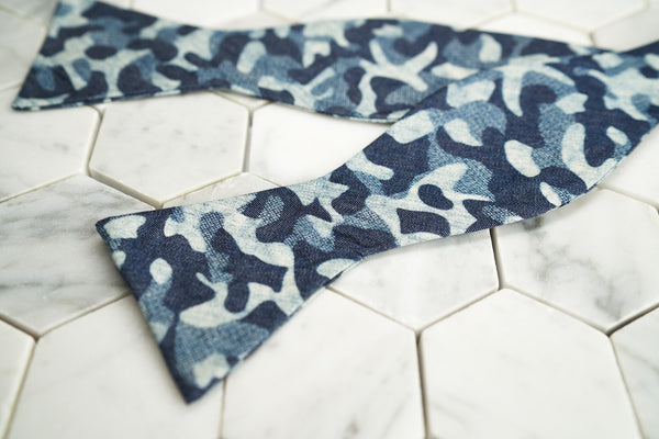 An image of an untied a blue indigo camouflage bow tie, laying across a white hex tile background.