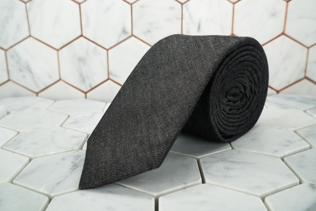 A black denim necktie made by Dear Martian, Brooklyn is shown rolled up and displayed against a hexagonal background.