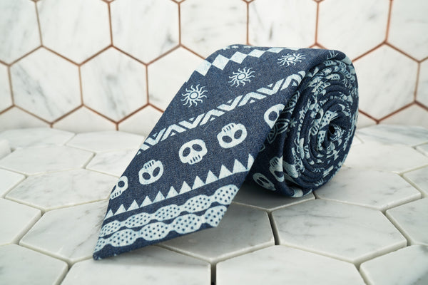 A product image of Dear Martian's Wallabout blue denim skull patterened necktie is rolled up against a white hexagonal background.